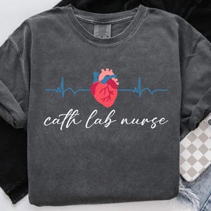 May include: Dark gray sweatshirt with a graphic of a red and pink anatomical heart, blue heartbeat line, and the words "cath lab nurse" in white script. The sweatshirt has a crew neck and long sleeves.