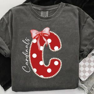 May include: Dark gray sweatshirt featuring a large red letter "C" with white polka dots and a red bow. The letter is outlined with pearls and the word "Cardinals" is written in white script. A phone with a checkered case is visible.