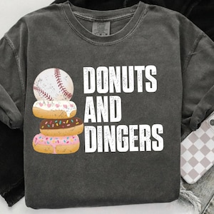 May include: Dark gray sweatshirt with the words "DONUTS AND DINGERS" in white. A baseball is stacked on top of three donuts with various toppings. The donuts are decorated with pink, brown, and white frosting.