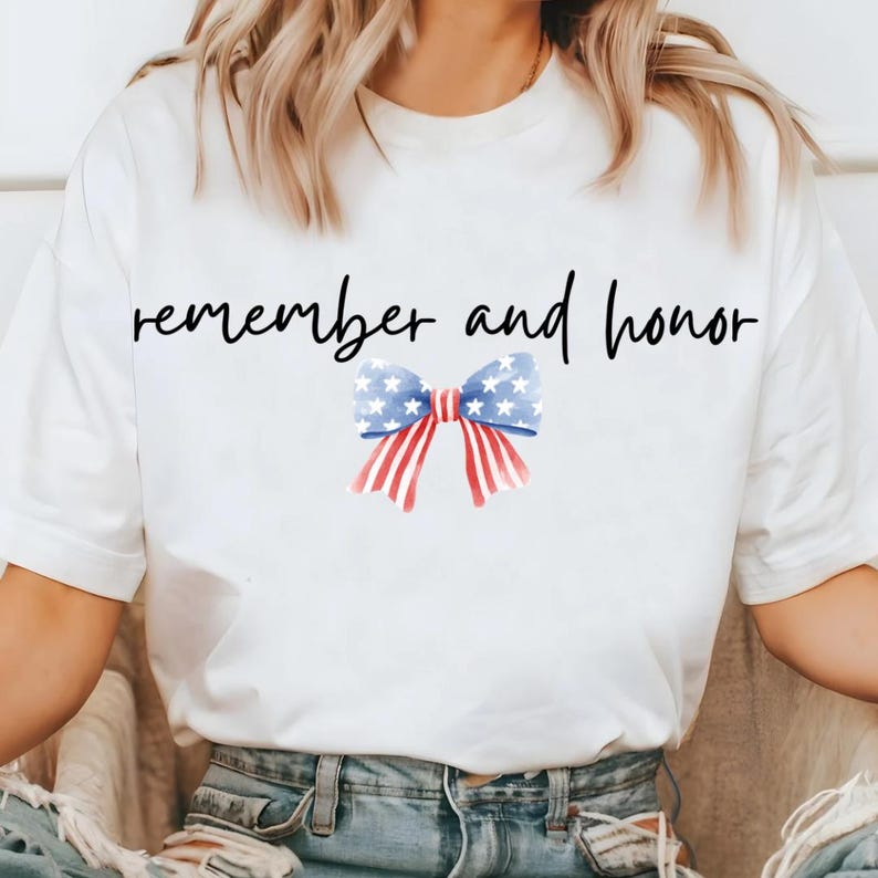 May include: White t-shirt with the words "remember and honor" in black script. A watercolor bow in red, white, and blue, with stars and stripes, is centered below the text. The shirt is a casual style.
