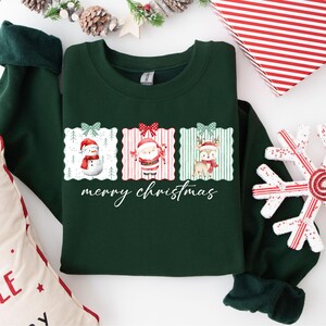 Puede incluir: Sudadera verde oscuro con un diseño festivo. El diseño presenta un muñeco de nieve, Papá Noel y un reno, cada uno dentro de un marco decorativo. Las palabras "merry christmas" están escritas en cursiva debajo de las imágenes. Un regalo de vacaciones perfecto.