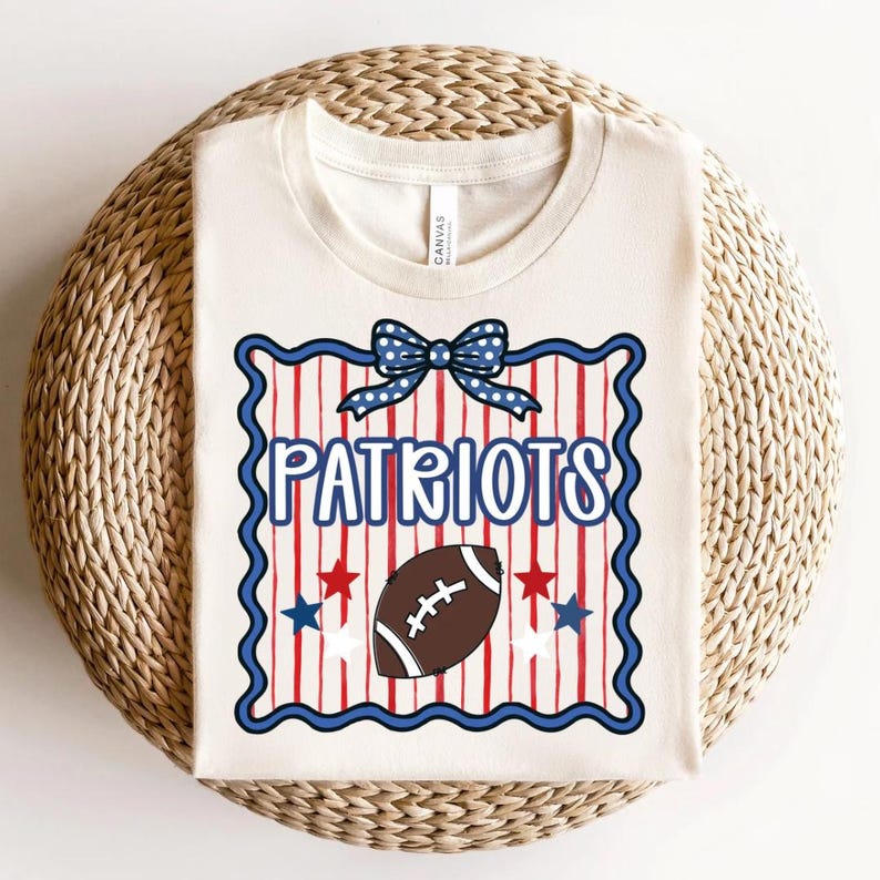 May include: A cream-coloured t-shirt featuring a graphic with a blue polka-dot bow at the top, the word "PATRIOTS" in blue letters, a brown football, and red and blue stars over a background of vertical red stripes inside a scalloped blue border.
