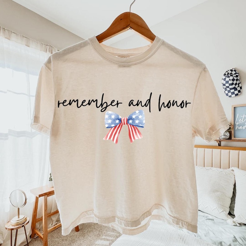 May include: A cream-colored t-shirt with the words "remember and honor" in black script. A patriotic bow graphic with blue stars and red stripes is centered below the text. The shirt is hanging on a wooden hanger.