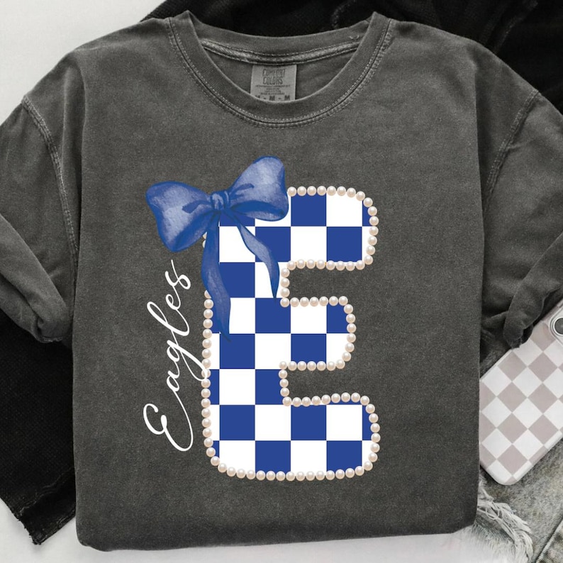 May include: Dark gray sweatshirt featuring a large, blue and white checkered letter "E" adorned with a blue bow and outlined with pearls. The word "Eagles" is written in script on the left side. The sweatshirt has a vintage, washed look.