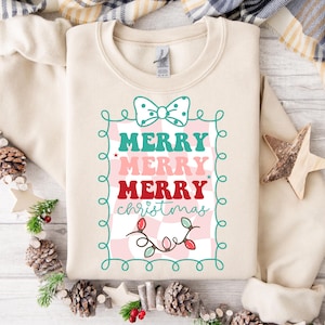 Puede incluir: Una sudadera crema con un diseño navideño festivo. El diseño presenta las palabras "MERRY MERRY MERRY Christmas" en verde azulado, rosa y rojo, enmarcadas por un borde verde azulado con un lazo y luces de cadena. La sudadera está rodeada de piñas y vegetación.