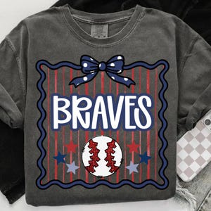 Puede incluir: Sudadera gris oscuro con un dise&ntilde;o de b&eacute;isbol. El dise&ntilde;o incluye la palabra "BRAVES" en blanco, una pelota de b&eacute;isbol, estrellas y un lazo. La pelota de b&eacute;isbol es roja y blanca, y las estrellas son rojas y azules.