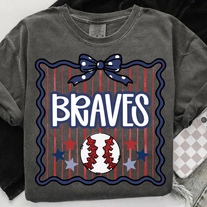 Puede incluir: Sudadera gris oscuro con un dise&ntilde;o de b&eacute;isbol. El dise&ntilde;o incluye la palabra "BRAVES" en blanco, una pelota de b&eacute;isbol, estrellas y un lazo. La pelota de b&eacute;isbol es roja y blanca, y las estrellas son rojas y azules.