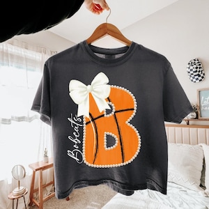 Könnte beinhalten: Ein anthrazitfarbenes T-Shirt mit einem großen orangefarbenen Basketball in Form des Buchstabens B, der mit weißen Perlen umrandet und mit einer weißen Schleife verziert ist. Das Wort Bobcats ist in weißer Schreibschrift links neben dem Motiv gedruckt.