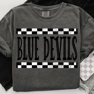 May include: A dark gray crew neck t-shirt with the text "BLUE DEVILS" in a bold, stylized font. The text is framed by black and white checkered patterns. The shirt has a vintage, worn look.