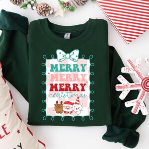 Puede incluir: Una sudadera verde oscuro con la frase "MERRY CHRISTMAS" en un marco decorativo. El texto es en verde azulado, rosa y rojo. Debajo del texto hay dibujos animados de un reno, Papá Noel y un oso polar. Un lazo blanco en la parte superior.