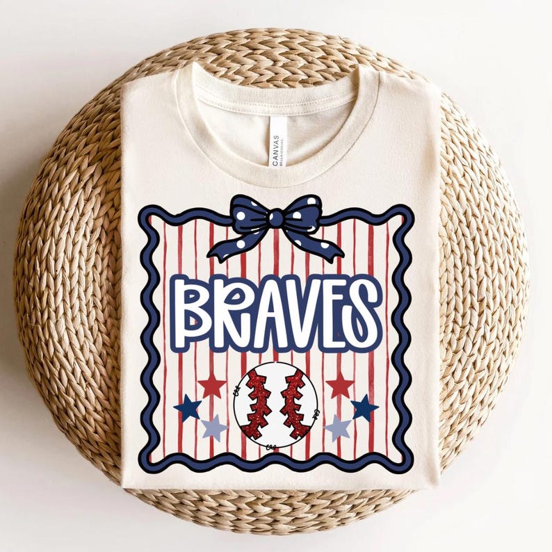 Puede incluir: Camiseta de color crema con un dise&ntilde;o tem&aacute;tico de b&eacute;isbol. El dise&ntilde;o incluye la palabra "BRAVES" en azul, una pelota de b&eacute;isbol, estrellas rojas y azules y un lazo. La camiseta est&aacute; doblada sobre una superficie tejida.