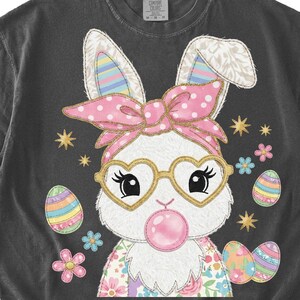 May include: Dark gray t-shirt featuring a cartoon bunny with heart-shaped glasses blowing a bubble. The bunny has a pink polka dot bow and is surrounded by Easter eggs and flowers. The shirt is a Comfort Colors brand.
