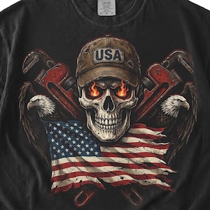 May include: Black t-shirt featuring a skull wearing a brown cap with "USA" text, fiery eyes, and crossed pipe wrenches. The design includes an American flag and eagle illustrations.