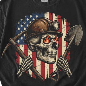 May include: Black t-shirt featuring a skull wearing a miner's helmet with a pickaxe and shovel crossed in front of an American flag design. The skull has fiery eyes. The shirt is a Comfort Colors brand.