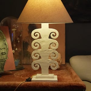 May include: A white table lamp with a brown lampshade. The lamp base has a distinctive, scroll-like design. The lamp is on a red table.