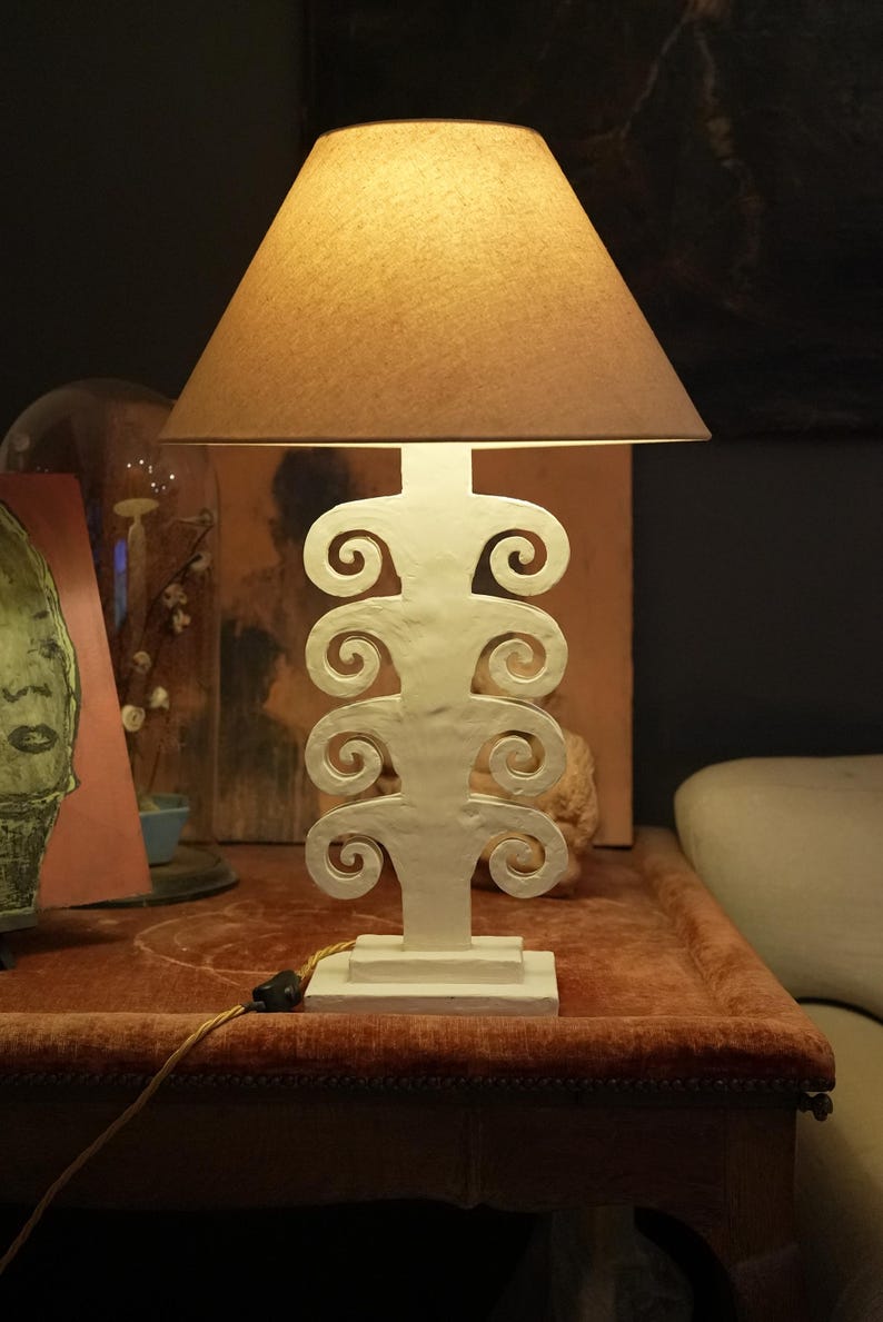 May include: A white table lamp with a brown lampshade. The lamp base has a distinctive, scroll-like design. The lamp is on a red table.