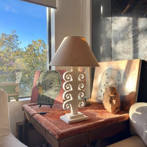 May include: A cream table lamp with a beige lampshade stands on a small table with a red velvet top. The lamp has a white, openwork base with a decorative design. Artwork and a sculpture are also on the table.