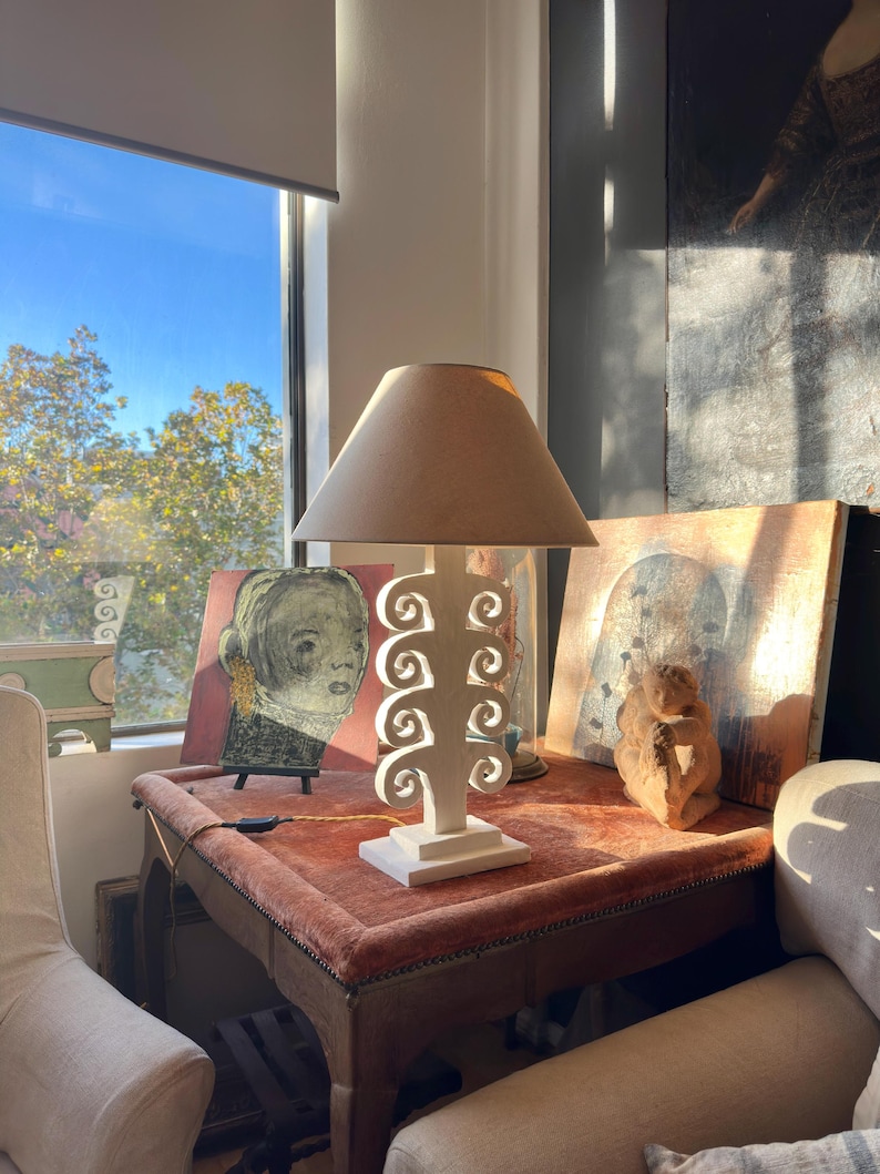 May include: A cream table lamp with a beige lampshade stands on a small table with a red velvet top. The lamp has a white, openwork base with a decorative design. Artwork and a sculpture are also on the table.