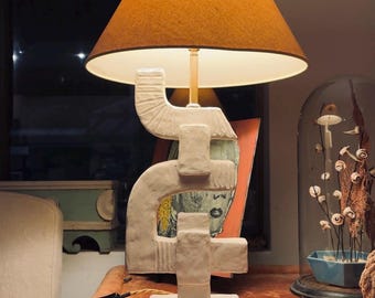 May include: A table lamp with a textured white base featuring stacked geometric shapes and a conical orange fabric lampshade. The lamp has a square base and a black power cord, emitting a warm light from beneath the shade.