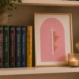 May include: A framed art print featuring a pink archway, a central sword illustration, and the text You Do Not Yield, displayed alongside a row of seven hardcover books by Sarah J. Maas, a small potted plant, a lit candle, and a classical bust.