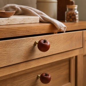 May include: Two round, dark brown wooden cabinet knobs shaped like apples, each featuring a small brown stem on top and a gold-toned metal base. These knobs are attached to the front of light brown oak wood drawers with a visible grain pattern.