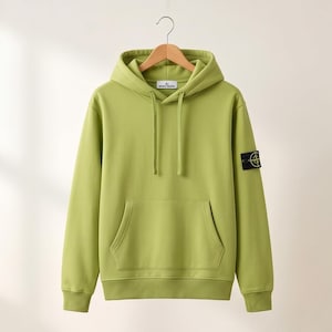 Puede incluir: Una sudadera con capucha verde claro con bolsillo tipo canguro y capucha con cordón. La sudadera tiene mangas largas y un pequeño parche negro con un logotipo en la manga izquierda. Colgada en una percha de madera.