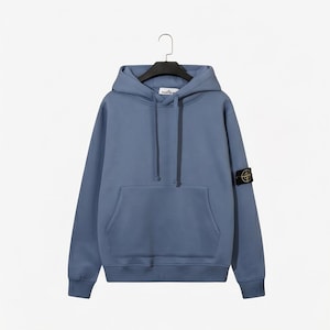 Puede incluir: Sudadera azul con capucha, bolsillo tipo canguro y un pequeño parche con el logotipo en la manga izquierda. La sudadera está colgada de una percha negra sobre un fondo blanco.