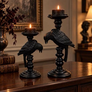 May include: Two black candle holders featuring sculpted raven figures perched on tiered pedestals. Each holder supports a small, dark, lit pillar candle. The raven designs are detailed with textured feathers and downward-facing heads.