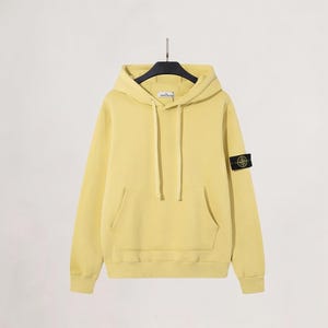 Puede incluir: Una sudadera con capucha de color amarillo pálido con capucha con cordón y bolsillo de canguro. La sudadera de manga larga tiene un pequeño parche en la manga izquierda. La sudadera con capucha está colgada en una percha negra sobre un fondo blanco.