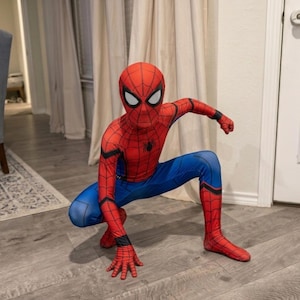 May include: A child wearing a Spider-Man costume, featuring a red and blue color scheme. The costume includes a red mask with white eye lenses, a red top with a black spider emblem, and blue pants. The child is crouching on a wooden floor.