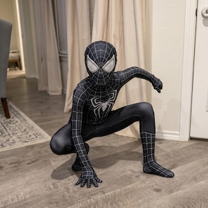 May include: A black and white Spider-Man costume. The costume features a spider emblem on the chest and a web pattern. The mask has large, silver-colored eye lenses. The person is crouching down on a wooden floor.