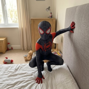 May include: A black and red Spider-Man costume with a mask, featuring a spider emblem on the chest. The costume has red accents on the shoulders and hands. The person is crouching on a bed with white sheets.
