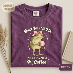 May include: A berry-coloured crew neck t-shirt featuring a graphic of a toad wearing a pink bow and holding a pink coffee mug. The text reads Don't Talk To Me Until I've Had My Coffee, surrounded by small pink hearts and white star accents.