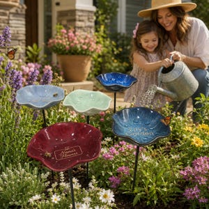 May include: Four flower-shaped garden stakes with glossy finishes in light blue, pale green, deep red, and dark blue. The red stake features the text Nana's Flowers, and the dark blue stake features the text Lily's Garden, both mounted on thin black metal rods.