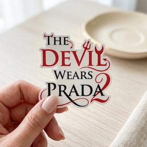 May include: A white sticker with the text "The Devil Wears Prada" in black and red. The word "Devil" is in red, with a trident symbol above it. The sticker is held by a hand.