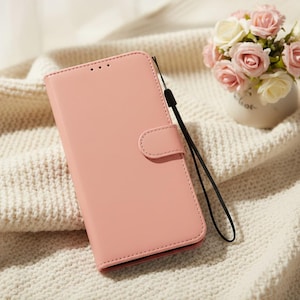 Puede incluir: Una funda de teléfono rosa claro con cierre magnético y correa de muñeca negra. La funda es de material liso y tiene un borde cosido. Un pequeño ramo de rosas rosas y blancas está en un jarrón blanco.