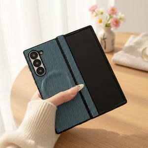 May include: A denim-effect phone case with a black frame and a triple-lens camera. The case is folded open, showing a black screen. The case is held by a person wearing a cream-coloured sweater.