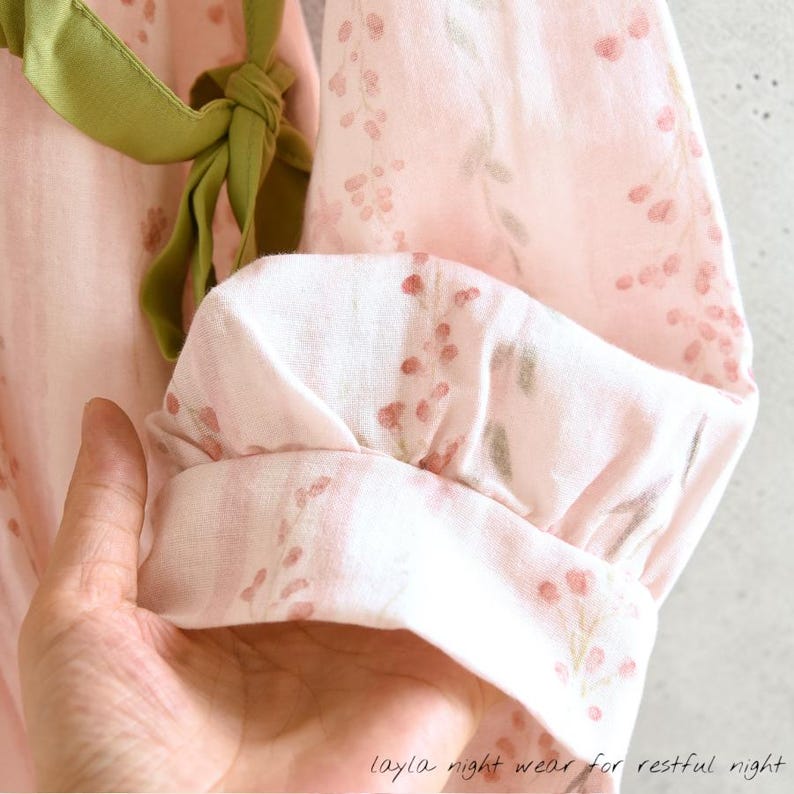 May include: Close-up of a pink floral-patterned garment with a green ribbon tie. The fabric features a delicate print of small red and green floral elements. The garment's sleeve is cuffed, and the text "layla night wear for restful night" is visible.