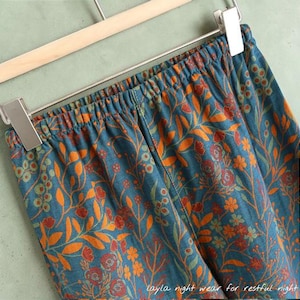 May include: Teal-colored pajama pants with an orange floral pattern, hanging on a wooden hanger. The elastic waistband is visible. The text "Layla night wear for restful night" is at the bottom.
