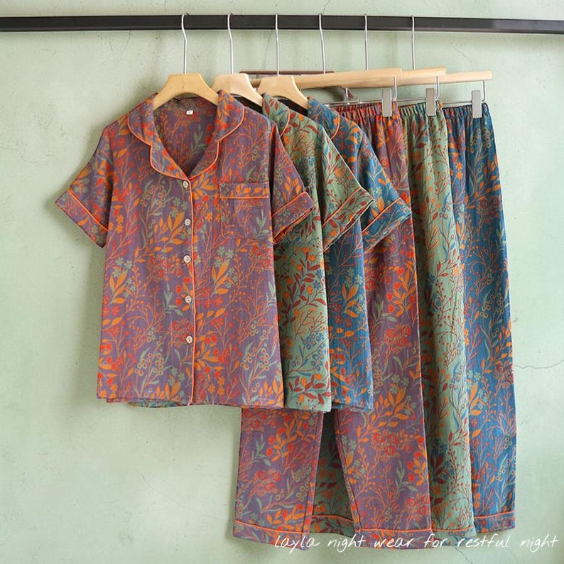 May include: A set of floral-patterned pajamas in shades of purple, orange, and green, hanging on wooden hangers. The short-sleeved top has a button-down front and a notched collar. The matching pants are long. The text "Layla night wear for restful night" is at the bottom.
