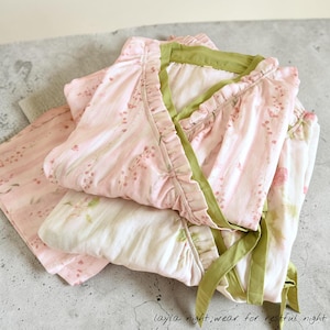 May include: A set of pink floral pajamas with green trim and ruffles. The pajama top has a wrap-around design with a tie closure. The matching pants are folded neatly beside the top. The text "layla night.wear for restful night" is visible.