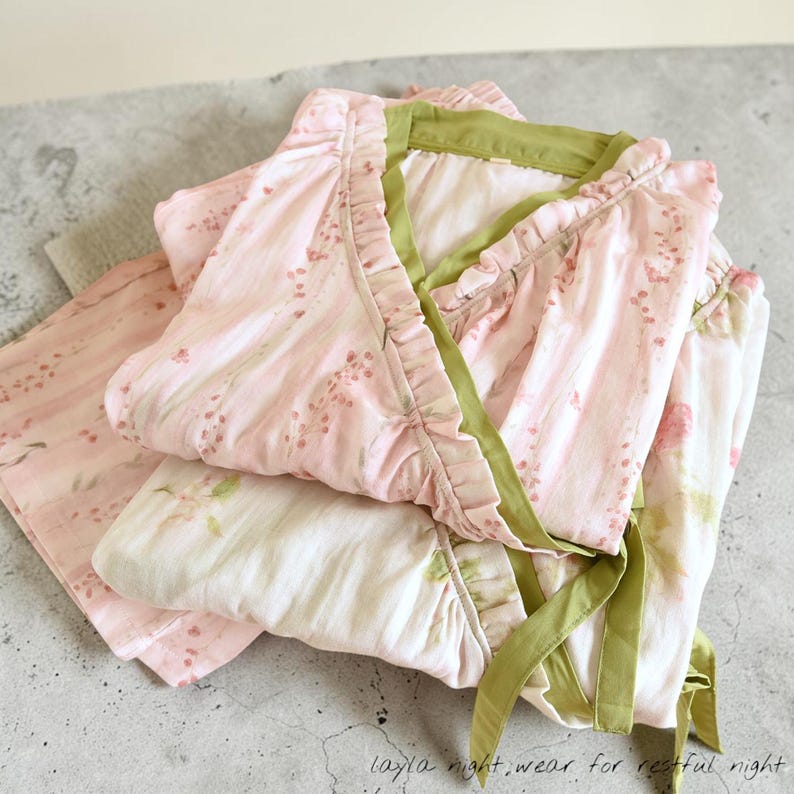 May include: A set of pink floral pajamas with green trim and ruffles. The pajama top has a wrap-around design with a tie closure. The matching pants are folded neatly beside the top. The text "layla night.wear for restful night" is visible.