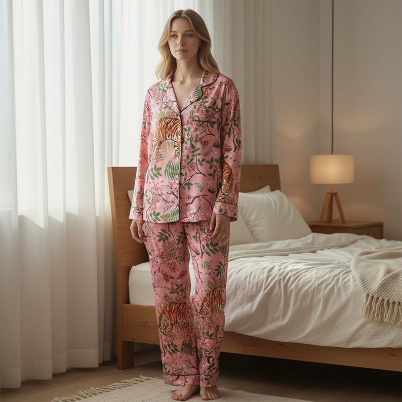 May include: Pink pajama set with a tiger and floral print. The long-sleeved top has a button-down front and black trim. The matching pants have a relaxed fit. The pajamas are made of a soft, silky material.