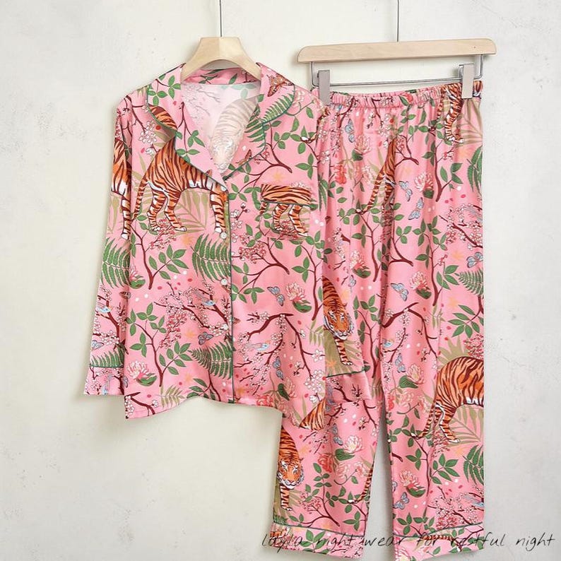 May include: Pink pajama set with a tiger and floral print. The long-sleeved top has a button-down front and a notched collar. The pants have an elastic waistband. The set features a repeating pattern of tigers, flowers, and leaves.