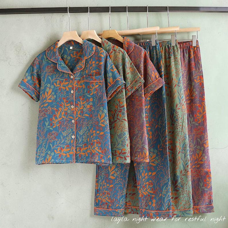 May include: Several sets of floral-patterned pajamas hang on wooden hangers. The short-sleeved tops and pants feature a blue background with orange and green botanical designs. The pajamas have orange trim and are made of a soft fabric.