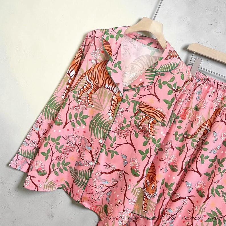 May include: Pink pajama set with a tiger and floral print. The long-sleeved top has a button-down front and a notched collar. The matching shorts have an elastic waistband. The design includes tigers, leaves, flowers, and butterflies.