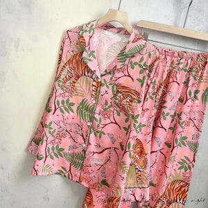 May include: Pink pajama set with a tiger and floral print. The long-sleeved top and pants feature a repeating pattern of tigers, butterflies, and foliage. The set has green trim and is displayed on a wooden hanger.
