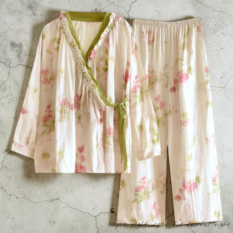 May include: A two-piece pajama set featuring a floral pattern in pink and green on a white background. The top has a wrap-around design with a green trim and tie. The matching pants are wide-legged. The text at the bottom reads "layla night wear, for restful night."