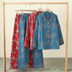Könnte beinhalten: Drei Pyjama-Sets hängen an einem Holzgestell. Ein Set ist rot mit einem Baummuster, und zwei Sets sind blau mit einem ähnlichen Design. Das blaue Pyjama-Oberteil hat einen Kragen und Knöpfe. Der Text "layla night wear for restful night" steht unten.