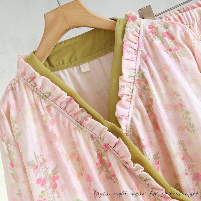 May include: Pink floral-patterned sleepwear set with ruffled edges and a green trim. The set includes a long-sleeved top and pants, hanging on a wooden hanger. The text "layla night wear for restful night" is visible at the bottom.